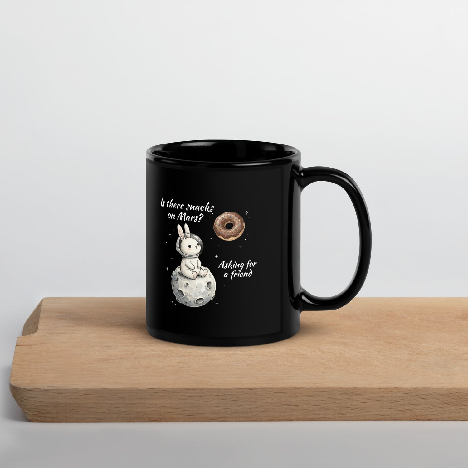 Black glossy ceramic coffee mug featuring a cute white astronaut bunny sitting on a moon looking at a floating chocolate donut with text Is there snacks on Mars? Asking for a friend